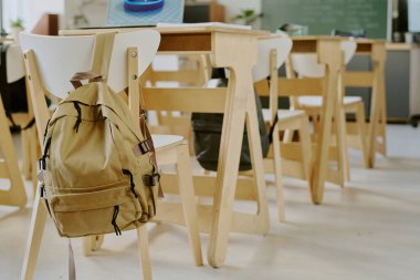 Sınıfın içi boş sandalye ve masalarla dolu, bir sandalyenin arkasında sırt çantasıyla, öğrenci yokluğu veya ilerleme dersi olduğunu gösteriyor.