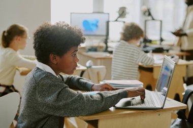 Ders sırasında sınıfta ders çalışmaya odaklanmış bir öğrenci ödevi tamamlamak için dizüstü bilgisayar kullanıyor. Arka planda emsallerle dolu sınıf ve eğitim materyalleriyle dolu