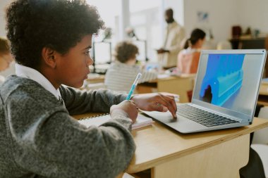 Modern sınıf ortamında, sınıf arkadaşları ve öğretmenlerle dolu bir laptopta eğitim aktivitesi üzerine çalışan genç bir öğrenci öğrenim faaliyetleriyle meşgul.