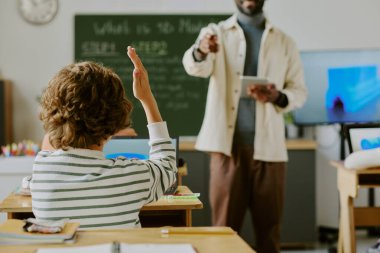 Sınıf ortamında öğrencinin elini kaldırmasına işaret eden tableti tutan öğretmen. Arkaplanda öğrencinin aktif olarak katıldığı karatahta ve projektör