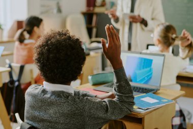 Öğretmen dersi açıklarken öğrenci el kaldırıyor. Arka planda öğretmenle iletişim kuran ve dizüstü bilgisayar kullanan diğer öğrenciler, canlı ve etkileşimli bir öğrenme ortamı öneriyorlar