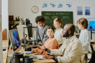 Öğretmen 3D modellik dersi yaparken öğrencileri kendine çekiyor. Çeşitli öğrencilerden oluşan bir grup bilgisayar ve 3D yazıcı ekipmanlarıyla sınıf ayarlarına odaklandı