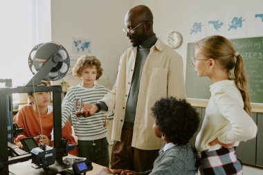 3D yazıcı ve eğitim materyalleri ile donatılmış bir sınıfta çeşitli öğrenci gruplarına 3D yazdırılmış nesneler gösteren Afro-Amerikan öğretmen