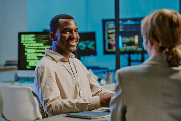 Cheerful young African American male cybersecurity specialist smiling while having conversation with colleagues during meeting