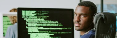 Genç Afrikalı Amerikalı erkek siber güvenlik uzmanı iş yerinde oturuyor gözlüklerini çıkarıyor ve kameraya bakıyor, web sitesi başlığı