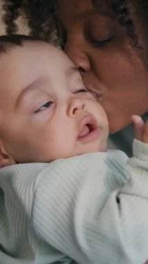 Dikey olarak gülümseyen, siyah kıvırcık saçlı, orta boylu, sevimli bir bebeği elinde tutan ve onunla vakit geçirirken oğlunu öpen Afro-Amerikan kadının fotoğrafı.