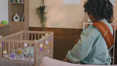 Genç Afrikalı Amerikalı bir kadın yatağında oturmuş bebek taşıyıcısını ya da doğum eşarbını göğsüne takıyor, kalkıyor, beşiğine yeni doğmuş bir bebekle çıkıyor ve oğlunu elleriyle tutuyor.