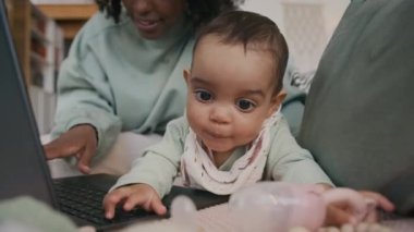Sevimli yeni doğmuş erkek bebek kanepede uzanıp dizüstü bilgisayarında klavye çalan ve sevimli oğluyla ilgilenen Afro-Amerikan bir kadının yanında oyuncakla oynuyor.