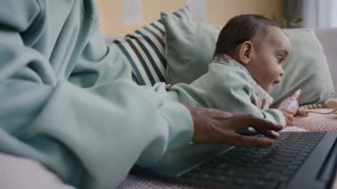 Genç Afro-Amerikan iş kadını, mavi uzun kollu sweatshirt 'lü. Dizüstü bilgisayarının tuşlarına basıyor. Kanepede tatlı oğlunun yanında oturmuş oyuncağıyla oynuyor.