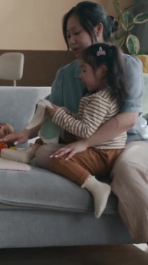 Dikey çekimde, annesinin yanında oturan ve el yapımı yumuşak oyuncak tavşanı sevimli yeni doğmuş erkek bebeğin yanına koyan Asyalı bir kız resmi.