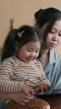 Genç Asyalı bir kadının dizlerinin üstüne çöküp, kameranın önünde oturmuş klavyenin tuşlarına basan sevimli kıza baktığı dikey bir fotoğraf.