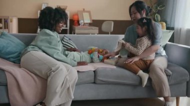 Annesinin yanında oturan, el yapımı yumuşak oyuncak tavşancığı sevimli yeni doğmuş erkek bebeğin yanına koyan sıradan giysili şirin Asyalı kızın yan görüntüsü.