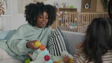 Uzun siyah saçlı küçük bir kız, gri kadife kanepede annesinin yanında mavi eşofmanlı Afro-Amerikan bir kadının önünde otururken tatlı yeni doğmuş bebeğine sarılıyor.