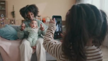 Cep telefonlu küçük bir kız dizlerinin üzerinde sevimli bir çocukla birlikte küçük bir Afrikalı Amerikalı kadının fotoğrafını çekiyor gri koltukta oturuyor ve sevimli bir fotoğrafçıya poz veriyor.