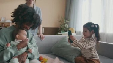 Mutlu genç Asyalı kadın akıllı telefonuyla küçük kızına yanaşıyor kanepeye oturuyor ve gülümseyen Afrikalı Amerikalı bekar annenin sevimli bebeğini kucağında tutarken fotoğrafını çekiyor.