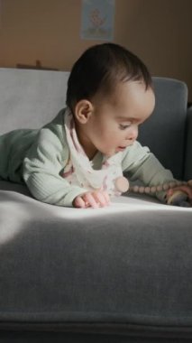 Açık mavi tulumlu, ev ortamında güneşli, kadife koltukta uzanmış, emzik ile oynayan ve yan bakan sevimli çocuğun dikey görüntüsü.