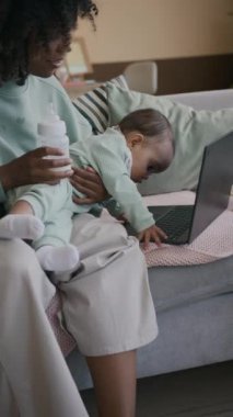 Dikey çekimde, dizüstü bilgisayarın yanındaki koltukta oturan ve bebek klavyeye dokunmaya çalışırken küçük oğlunu şişeden sütle besleyen gündelik giysili bir anne görülüyor.