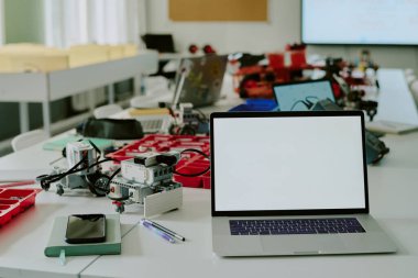 Orta boy dizüstü bilgisayar, beyaz ekran ve robotların bir kısmı boş sınıftaki ahşap masanın üstünde.