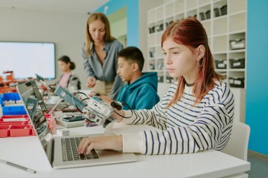 Okulda robotik dersinde robotların çalışma kapasitesini kontrol etmek için dizüstü bilgisayar kullanan çizgili tişörtlü, kızıl saçlı bir kız.