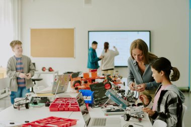 Esmer kız ve öğretmen birlikte tekerlek üzerinde robot inşa ederken diğer sınıf arkadaşları arka planda bulanık bir şekilde etkileşimli beyaz tahtaya yazı yazıyorlar.