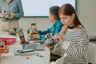 Kafkasyalı liseli kız masasında oturup robotunu tamir ederken sınıf arkadaşı da arka plandaki tanınmayan öğretmeni dikkatle dinliyor.