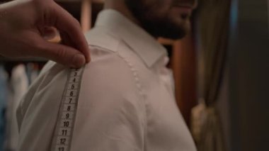 Beyaz gömlekli adamın omuz ölçüsünü ölçen tanınmayan terzinin elleri, soyunma odasında ona yardım ederken yakın çekim görüntüsü.