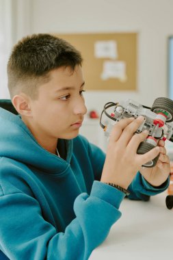 Mavi kapüşonlu bir çocuğun elinde tekerlekleri olan robotu tutup ona bakarken dikey görüntüsü.