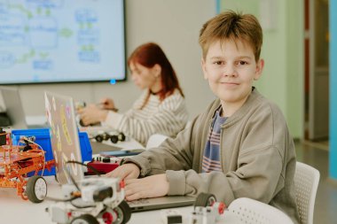 Sınıfta dizüstü bilgisayar kullanırken kameraya bakan çocuğun orta boy yakın portresi.