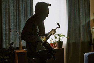 Siyahlı Afro-Amerikan hırsızın kirli deri eldivenler giydiği ve elinde metal levyeyle apartman dairesini gezerken yan görüntüsü.