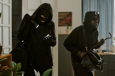 Afro-Amerikan suç ortağı elinde levyeyle odayı görürken, kar maskeli tanınmayan bir hırsızın orta ölçekli görüntüsü.