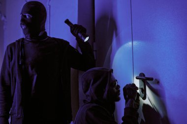 Kar maskeli bir hırsız kapıyı aydınlatıyor ve suç ortağı kilidi açarken etrafa bakıyor.