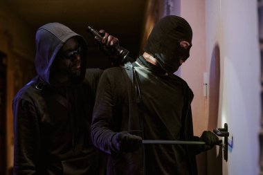 Kapı kilidini aydınlatan fenerli bir hırsız. Bu arada kar maskeli ortağı kilidi açmak için levye kullanıyor.