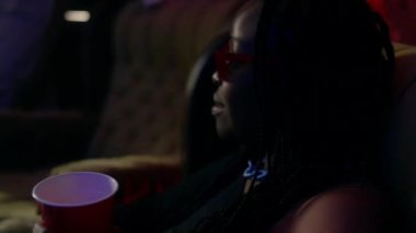 Siyah kızın plastik bardakta oturduğu ve neşeli beyaz adamın onun yanındaki koltuğa oturduğu, sonra da yanında duran bayan arkadaşına bir şişe bira verdiği fotoğraf.