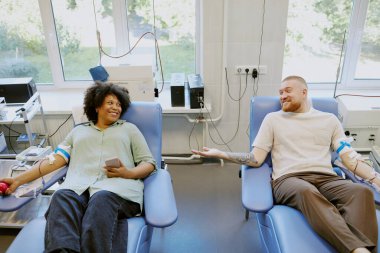 Tıbbi sandalyelerde oturan iki kişi kan bağışlıyor, birbirleriyle nişanlanıyorlar. Çeşitli tıbbi ekipmanlar görünür, klinik bir ortam yaratıyor.