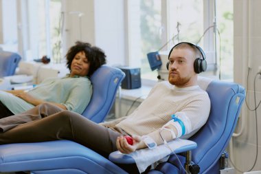 Kulaklık takan bir adam ve sandalyelerin oraya yaslanmış bir kadın. Rahat ve rahat görünürken modern kliniğe bağışta bulunmak.