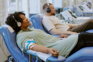 Afrikalı Amerikalı bir insan, arka plandaki diğer hastalarla müzik dinlerken kan bağışı yapıyor. Kolunda serum olan bir koltukta oturuyor.