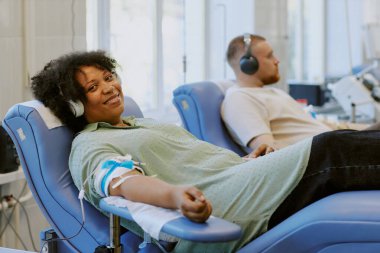 Afro-Amerikalı kadın ve beyaz adam koltuklarda dinlenirken kan bağışlıyorlar. Parlak bir tıbbi tesiste kan verirken kulaklıkları dinliyorlar.