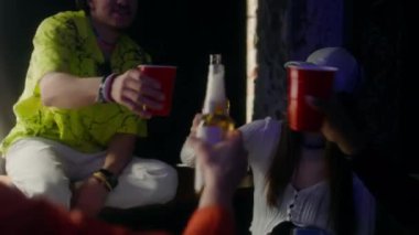 Yakın çekimde, elinde bira şişesi olan genç bir adam partide kutlama yaparken üç arkadaşıyla kadeh kaldırıyor.