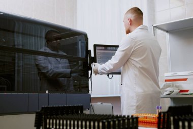 Steril laboratuvar ortamında çalışan bilim adamı, bilgisayar ekranındaki verileri incelerken aynı zamanda temiz ve düzenli çalışma alanlarındaki gelişmiş laboratuvar araçlarını da inceliyor.