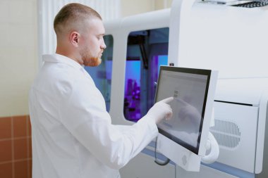 Laboratuvar önlüklü erkek bilim adamı modern laboratuvar ortamında dokunmatik ekran monitörü üzerinde inceleme yapıyor, araştırma ve bilimsel analiz yürütüyor.