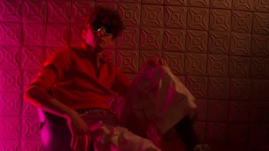 Kulüpte sandalyeye oturmuş arka planda beyaz fayanslar olan kameraya bakarken iki koluyla dizini kavrayan gözlüklü genç bir adamın portresi.