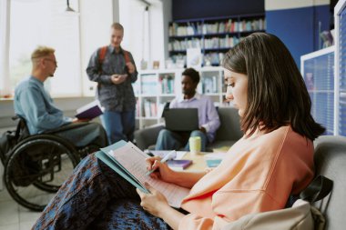 Bir grup öğrenci modern, parlak bir kütüphanede oturup ders çalışıyor, sohbet ediyorlar ve elektronik cihazlar kullanıyorlar. Çok ırklı bireyler kapsayıcı ortamlarda işbirliği içinde çalışırlar.