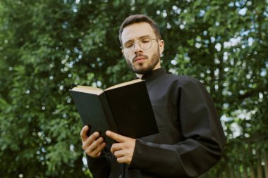 Kafkasyalı rahibin yuvarlak gözlükleriyle kilisenin avlusunda dinlenirken siyah örtülü İncil okurken alçak açılı çekimi.