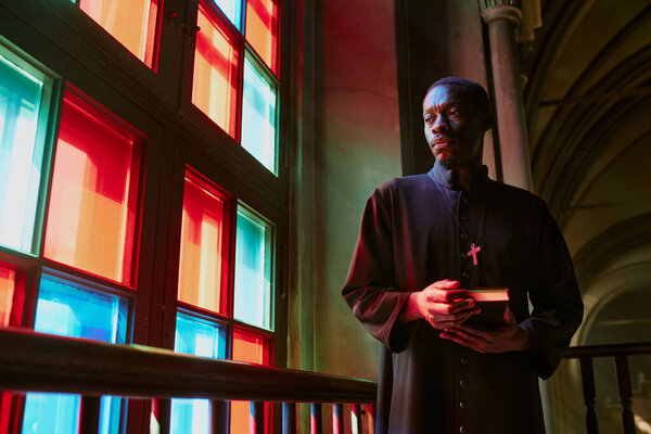 Low angle shot of African American priest with Bible looking out of multicolored stained window