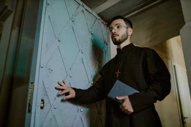 Siyah cüppeli beyaz papazın elinde ahşap haç ve elinde İncil olan ağır metal kapıyı açtığı alçak açılı bir kare.