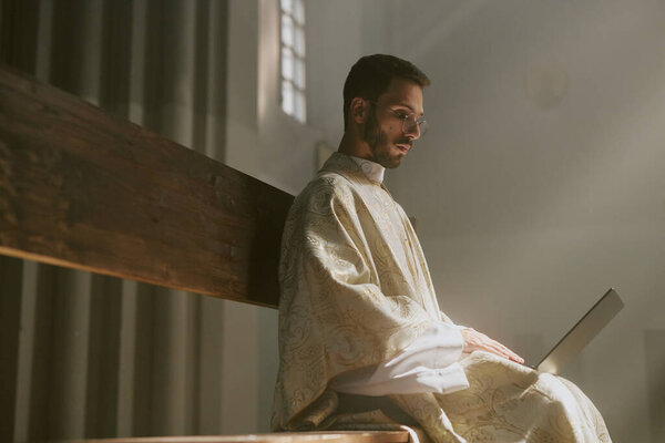 Young Caucasian priest dressed in golden white robe surfing Internet while sitting in church