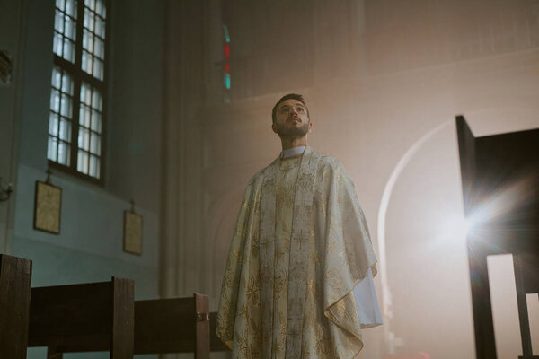 Low angle shot of young Caucasian priest dressed in white long robe with golden patterns looking at beautiful ceiling of church