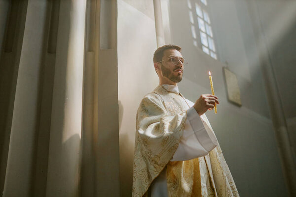 Low angle shot of ample church nave with young Caucasian priest in golden white robe holding long wax candle during service inside
