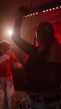 Pozitif Afrikalı Amerikalı bir kızın dans pistinde neon ışıkları ve flaşlarla dans ettiği dikey çekim. Arka planda dans eden iki genç adam.