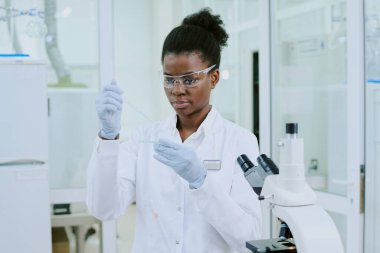 Pipet tutan Afrikalı Amerikalı kadın bilim adamı, güvenlik gözlüğü ve laboratuvar önlüğü takıyor, iyi donanımlı, parlak ışıklı bir laboratuarda deneye odaklanıyor.